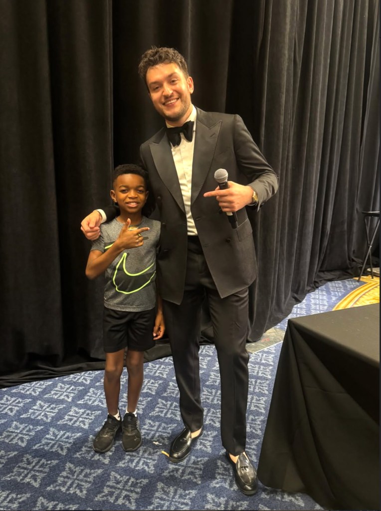A man in a suit holding a microphone, standing next to a smiling young boy in a gray t-shirt and black shorts.